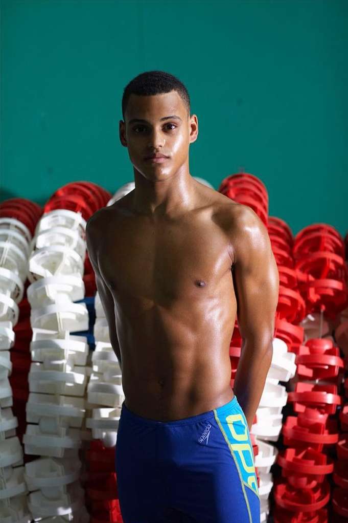 An athletic man with muscular build, posing shirtless in blue swimsuit against a green wall and competition swimming lane divider rack.