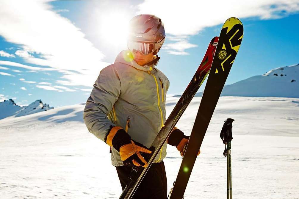 Person in ski gear holding skis on snow-covered mountain, wearing helmet and goggles, with sun shining in background, winter sport activity.