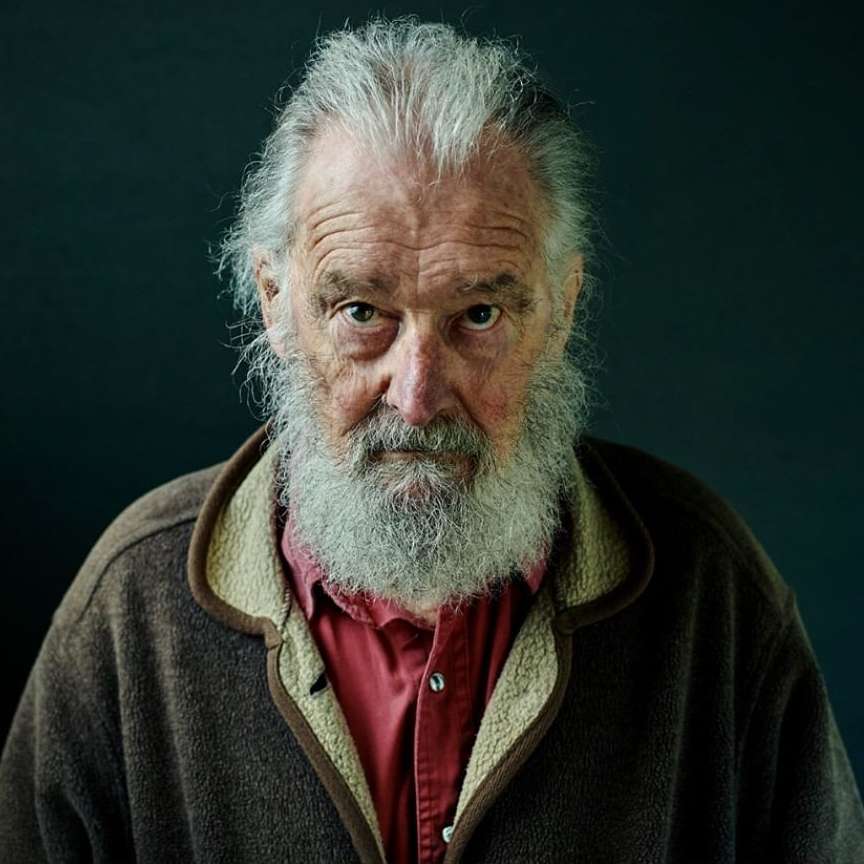 Portrait of an older man with a gray beard and hair wearing a brown jacket and red shirt against a dark green background.