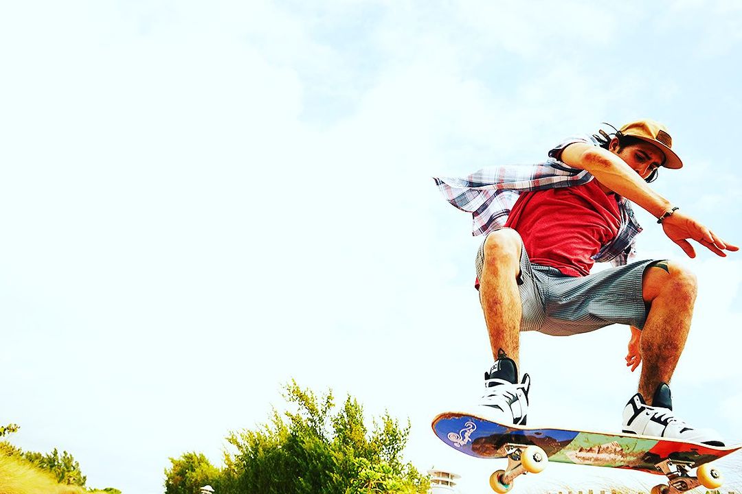 Male skateboarder in mid-air wearing casual clothes and hat, captured from a low angle with a bright blue sky backdrop.