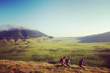 Three hikers with backpacks walking across a grassy hillside valley beneath a wide mountain landscape.