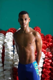 Shirtless young man in blue swim shorts standing before a stack of red and white lane markers in Los Angeles