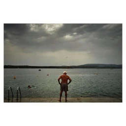 Elderly man standing with hands on hips on a concrete pier overlooking a calm body of water under a dark cloudy sky