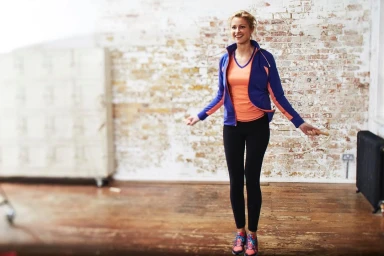 A smiling woman skipping rope indoors wearing a blue jacket and black leggings