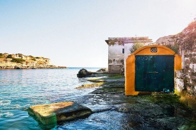 Coastal view with a vibrant yellow and green boathouse structure beside calm sea water under bright sunlight.