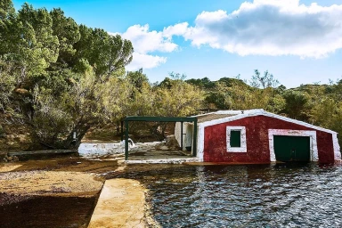 Red and white coastal boathouse at water edge near trees under blue sky with clouds