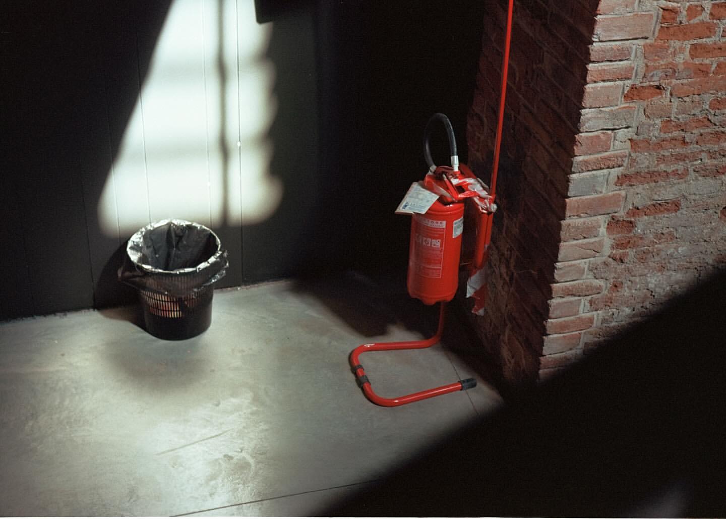 Indoor image featuring a red fire extinguisher beside a brick wall, contrasted with a black waste bin and light shadows on the floor.