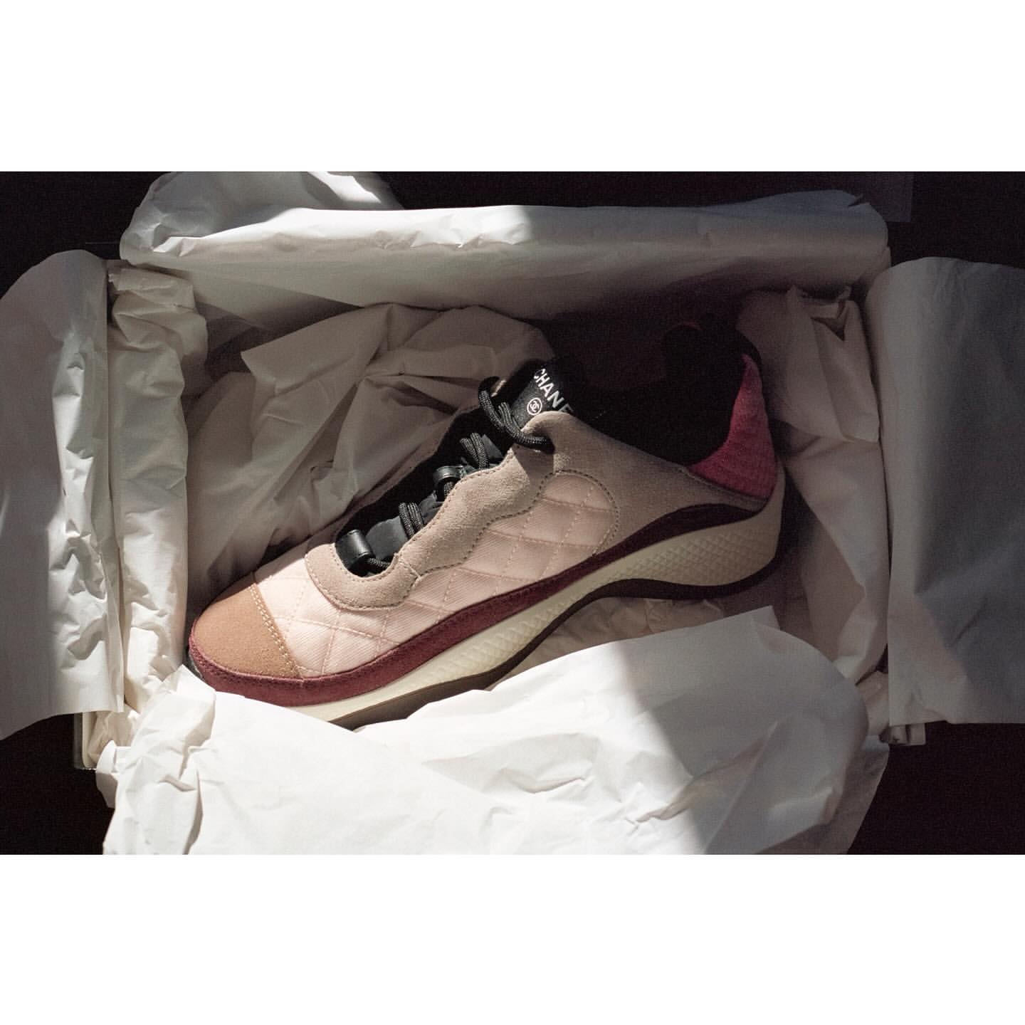 Fashionable pink and tan Chanel sneaker in a white box with crumpled tissue paper, lit by bright sunlight creating strong shadows.