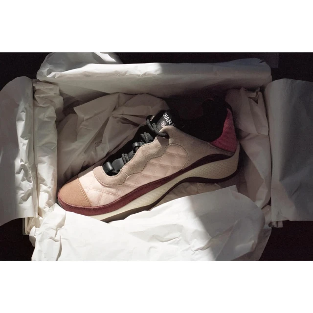 Fashionable pink and tan Chanel sneaker in a white box with crumpled tissue paper, lit by bright sunlight creating strong shadows.