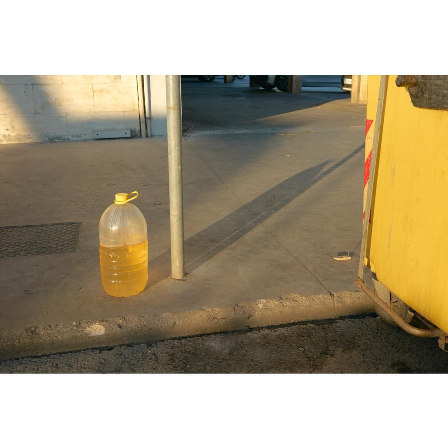 A large, partially-filled jug of yellow liquid sits outdoors on a concrete surface near a metal post and part of a yellow structure.