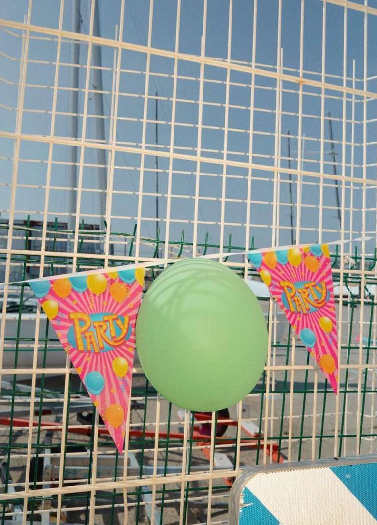 A green balloon and two party flags hang on a wire fence against a blurred background of blue sky and wire mesh, creating a festive scene.