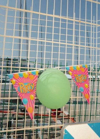 A green balloon and two party flags hang on a wire fence against a blurred background of blue sky and wire mesh, creating a festive scene.