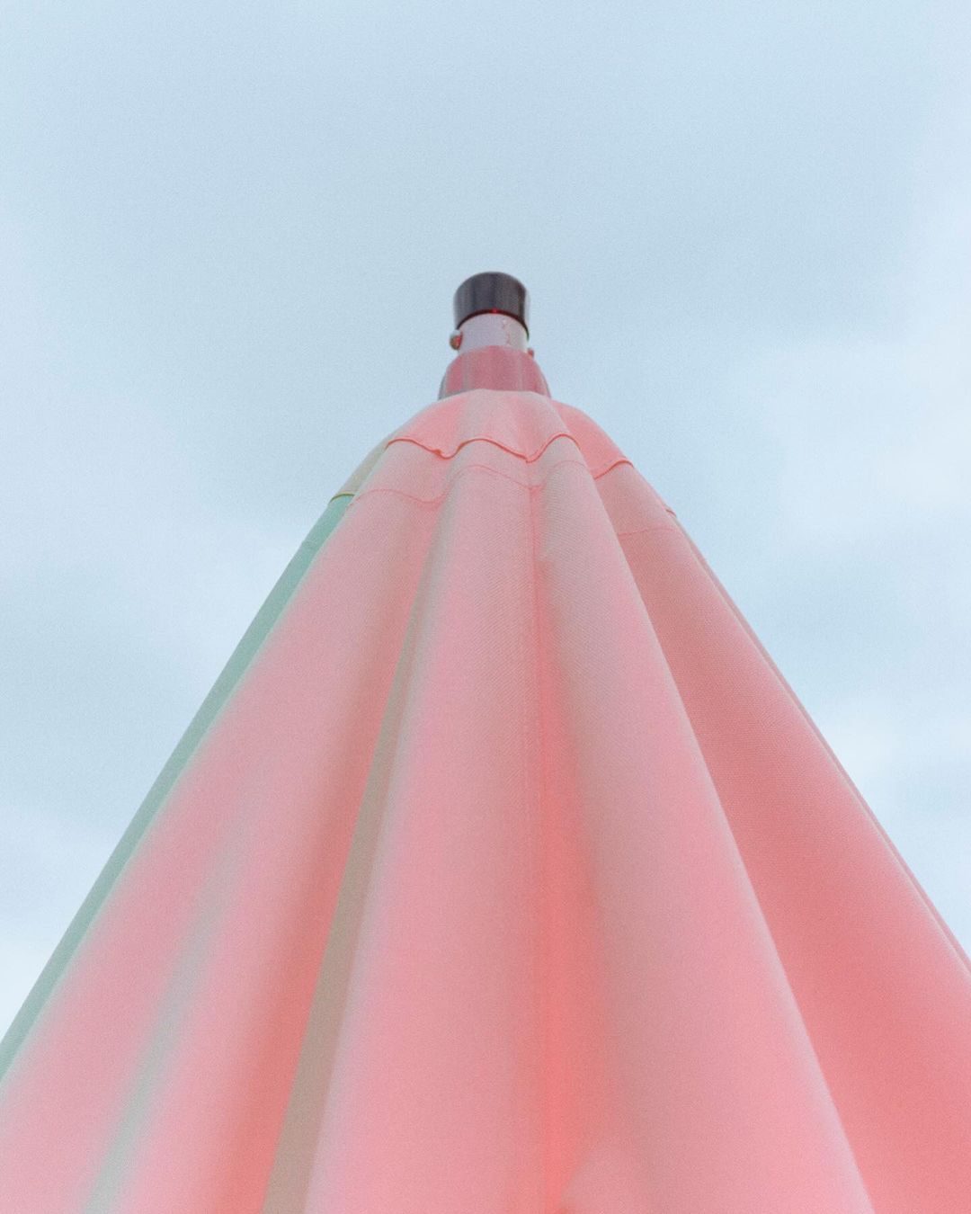 Low angle shot of a pink umbrella with metal pole against a light blue sky, creating a minimalist and colorful summer image.