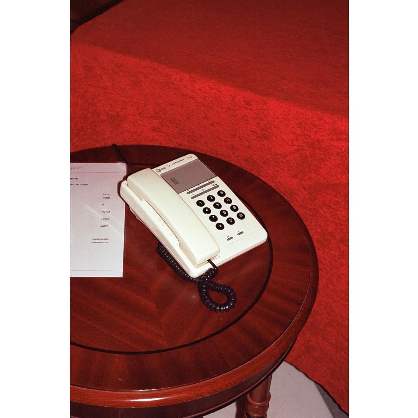 Vintage-style ivory-colored landline telephone with black cord rests on a round wood table with a red-hued bed in the background.