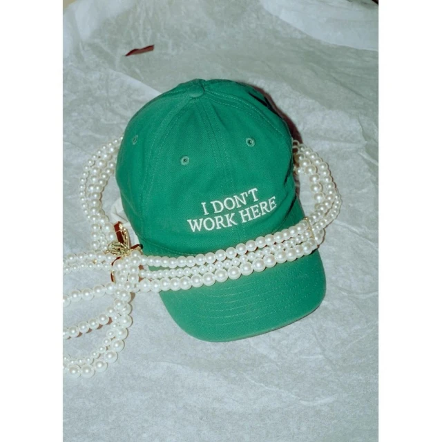 Green baseball cap with 'I DON'T WORK HERE' embroidery, styled with multiple strands of pearl necklaces on a white surface.