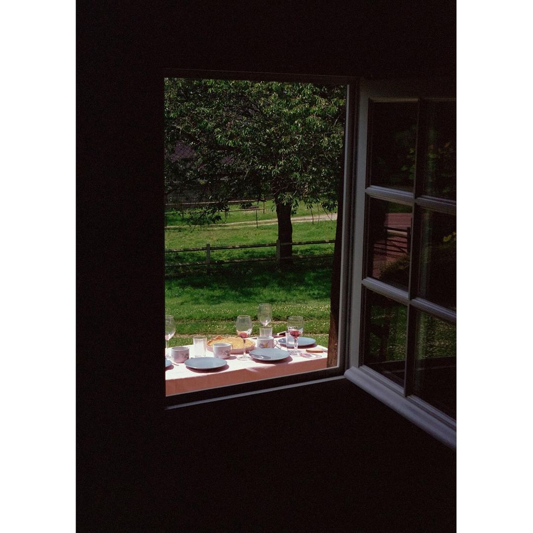 Table setting with plates, glasses, and utensils on a table seen through a window, with greenery and a rural landscape in the background.