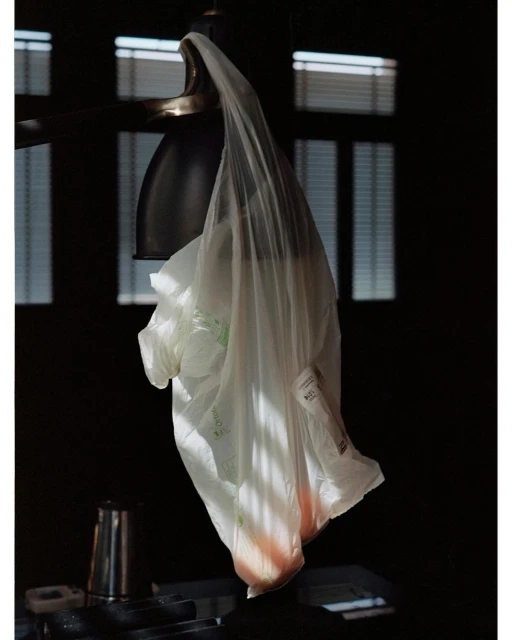 A plastic bag hangs from a light fixture indoors, bathed in partial sunlight, with blinds visible through a window in the background.