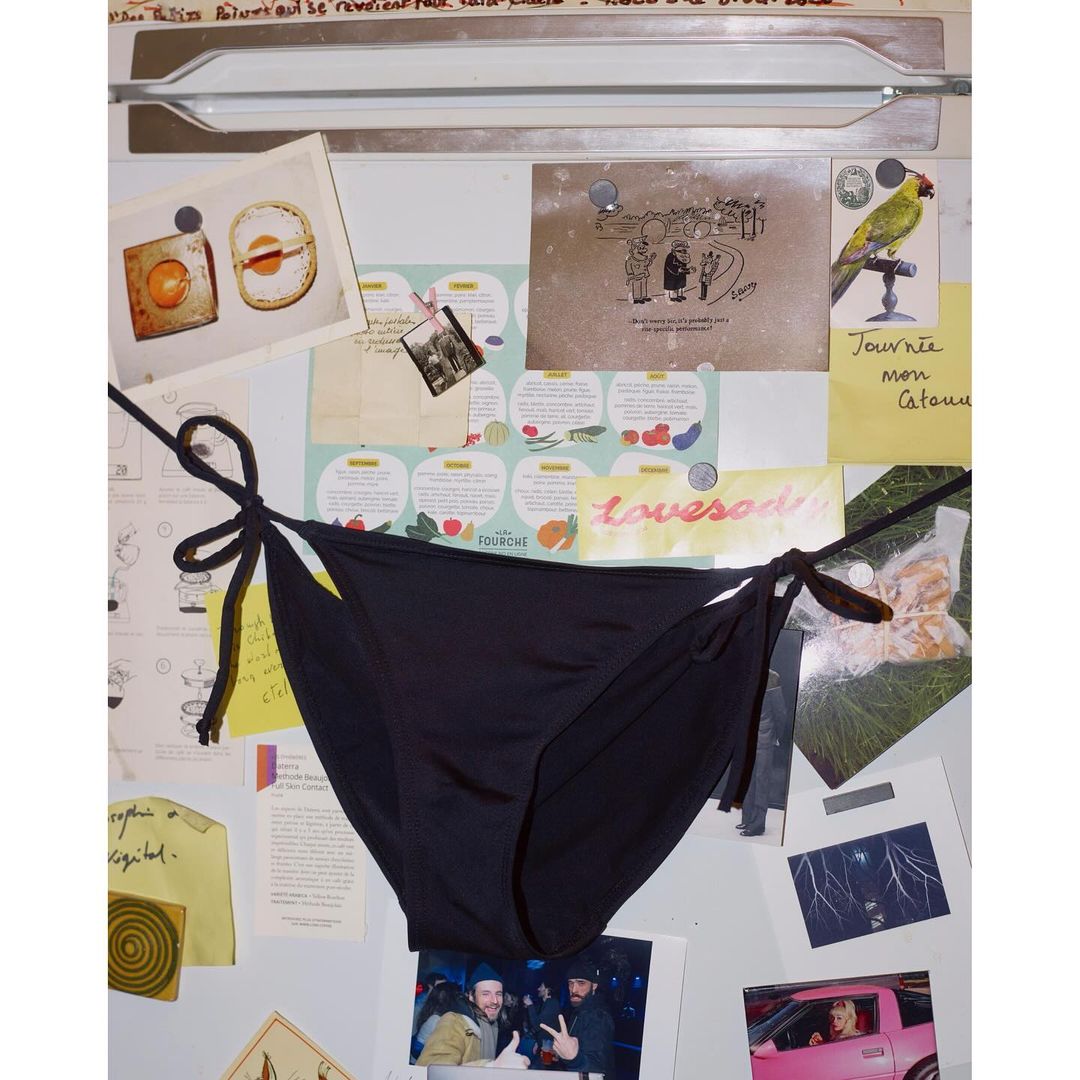 Black string bikini bottoms are centered in a studio shot against a background of various notes, photos, and papers on a metal surface.