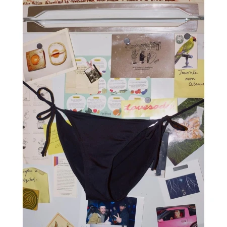 Black string bikini bottoms are centered in a studio shot against a background of various notes, photos, and papers on a metal surface.