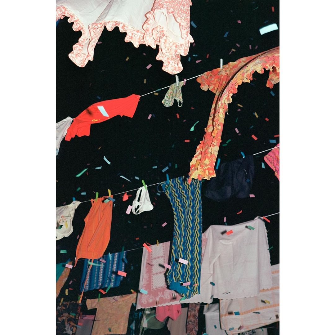 Colorful laundry hanging on clotheslines under confetti in a dark, outdoor setting. Garments in various styles, textures, and patterns.