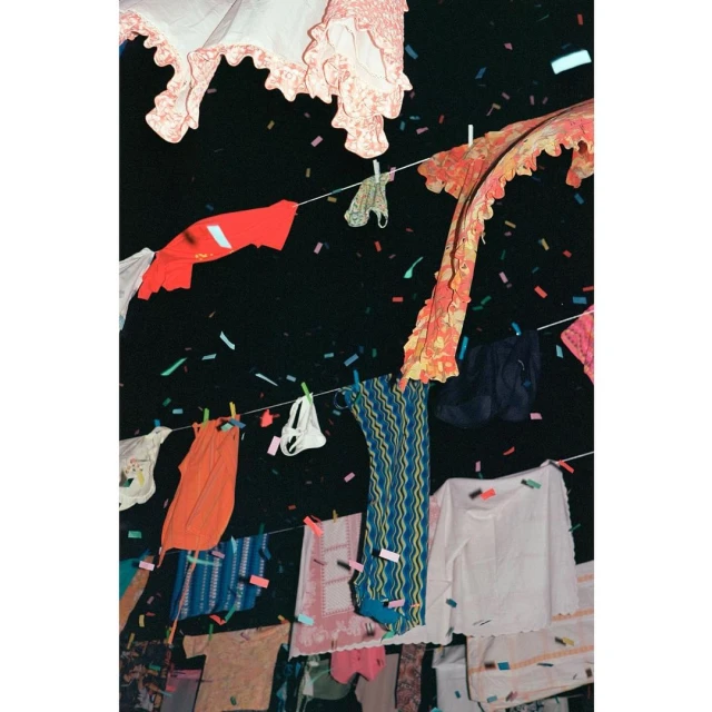 Colorful laundry hanging on clotheslines under confetti in a dark, outdoor setting. Garments in various styles, textures, and patterns.