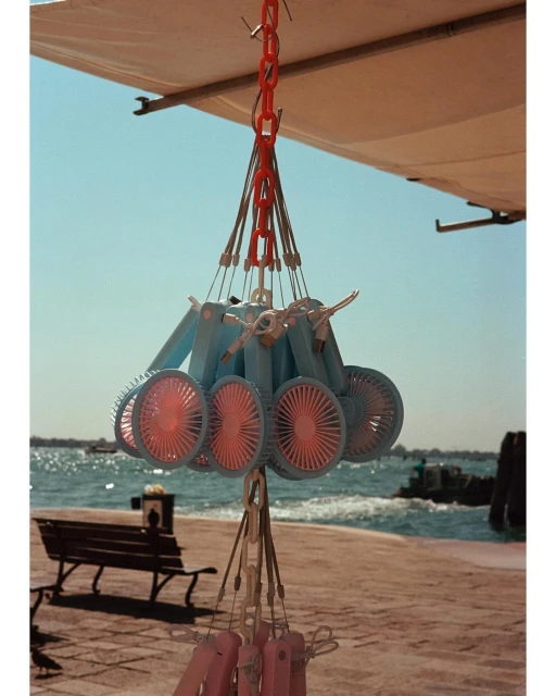Several blue and pink handheld fans hang from orange chain links against a bright blue sky, tan awning, and ocean backdrop.
