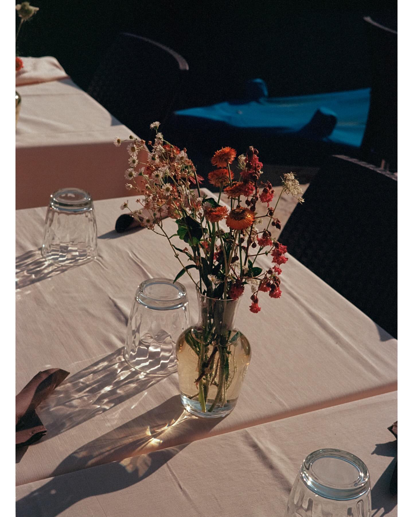 Still life photography features a flower arrangement in a vase on a table, accompanied by glassware, set in an indoor environment with natural lighting.