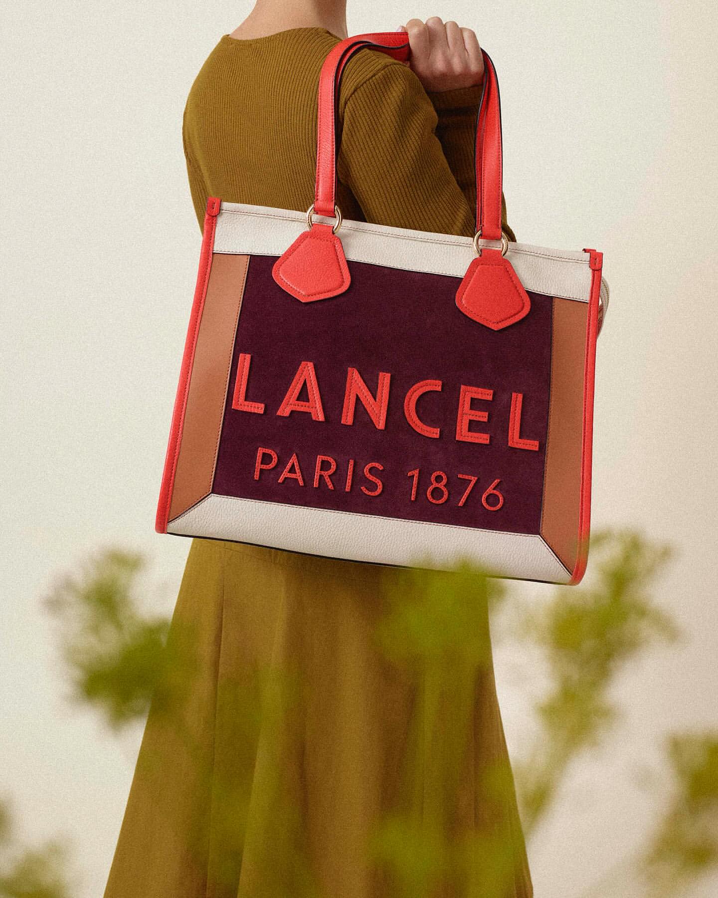 Lancel Paris 1876 burgundy tote bag with coral trim and matching handle held by a person wearing an olive green dress, studio shot.