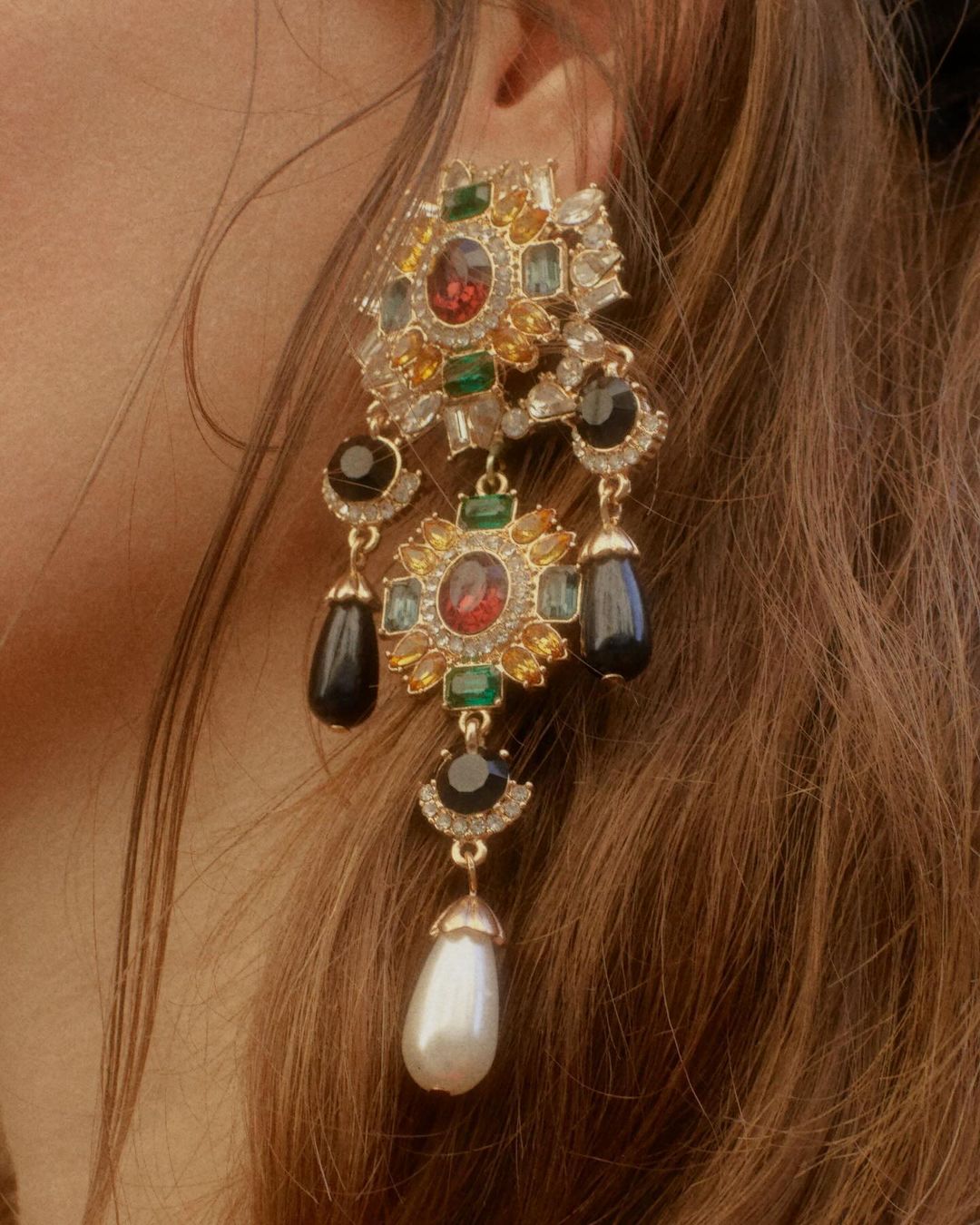 Close-up shot of a woman's ear with a gold chandelier earring featuring pearls, emeralds, rubies, and diamonds, showcasing jewelry and fashion.