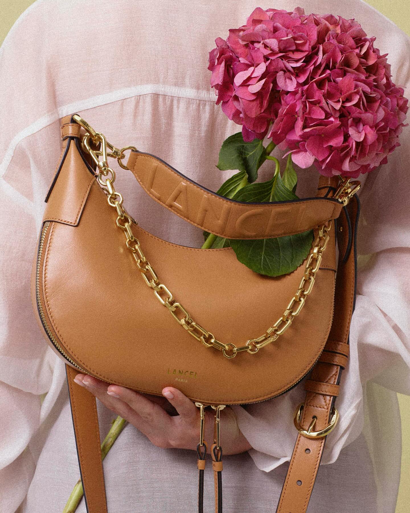 Tan Lancel handbag draped on person in white shirt with pink flowers. Gold chain and hardware details. Fashion product shot.
