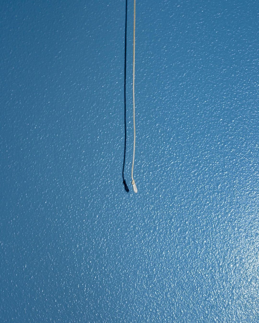 Close-up of a white string hanging against a blue wall, casting a shadow. Simple composition with a clean, textured backdrop.