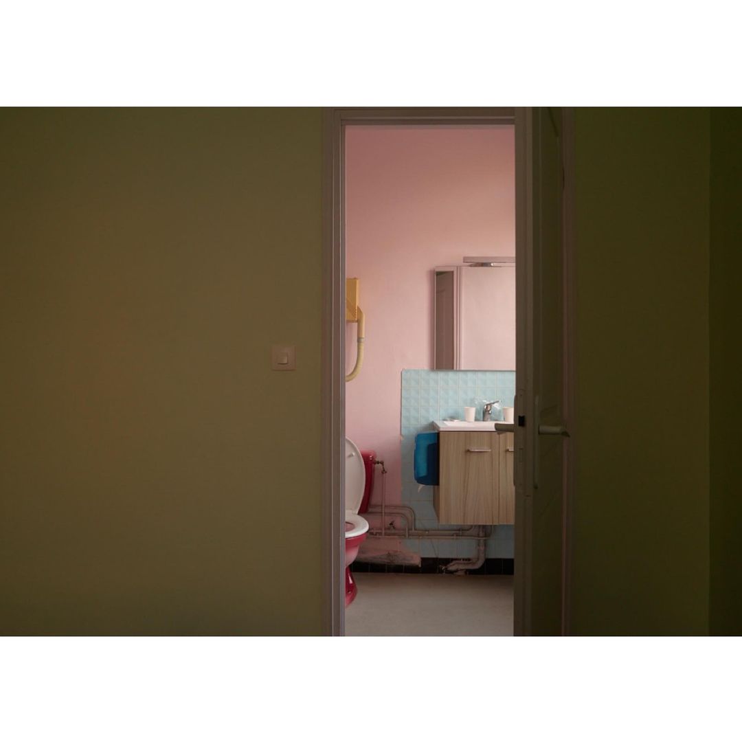 Interior shot of a bathroom visible through a doorway, featuring a sink, mirror, toilet, and green and pink walls with a vintage feel.