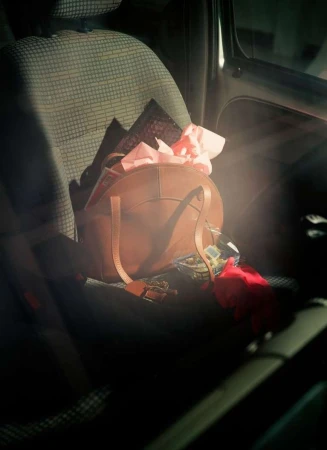Brown leather purse sits on a car seat, filled with pink tissue paper and other items, illuminated by sunlight in a dark interior.