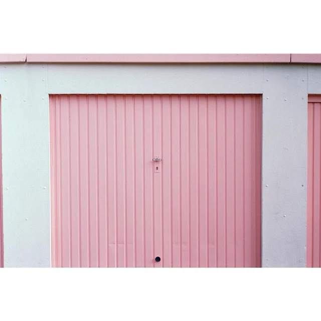 A symmetrical eye-level shot of a pink metal roll-up garage door with white trim on a building exterior, creating a minimalist facade.