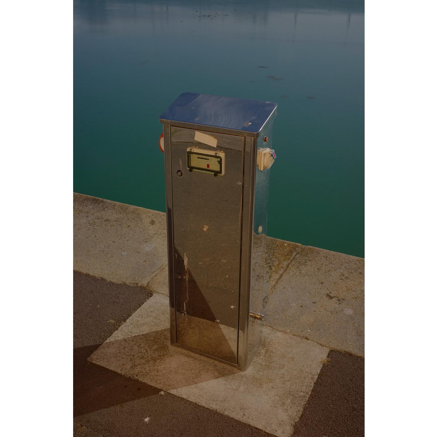 Stainless steel utility box sits on a paved waterfront with a calm sea in the background under an overcast daytime sky.
