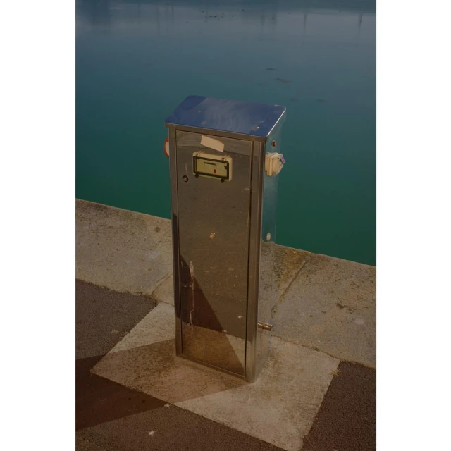Stainless steel utility box sits on a paved waterfront with a calm sea in the background under an overcast daytime sky.