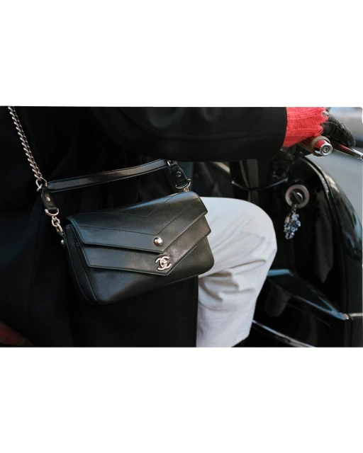 A black leather shoulder bag with a chain strap and the Chanel logo is slung across a person's body as they sit on a black moped.