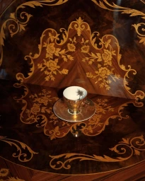 An ornate metallic gold teacup and matching saucer rest on a polished marquetry wood table with intricate floral scrollwork.