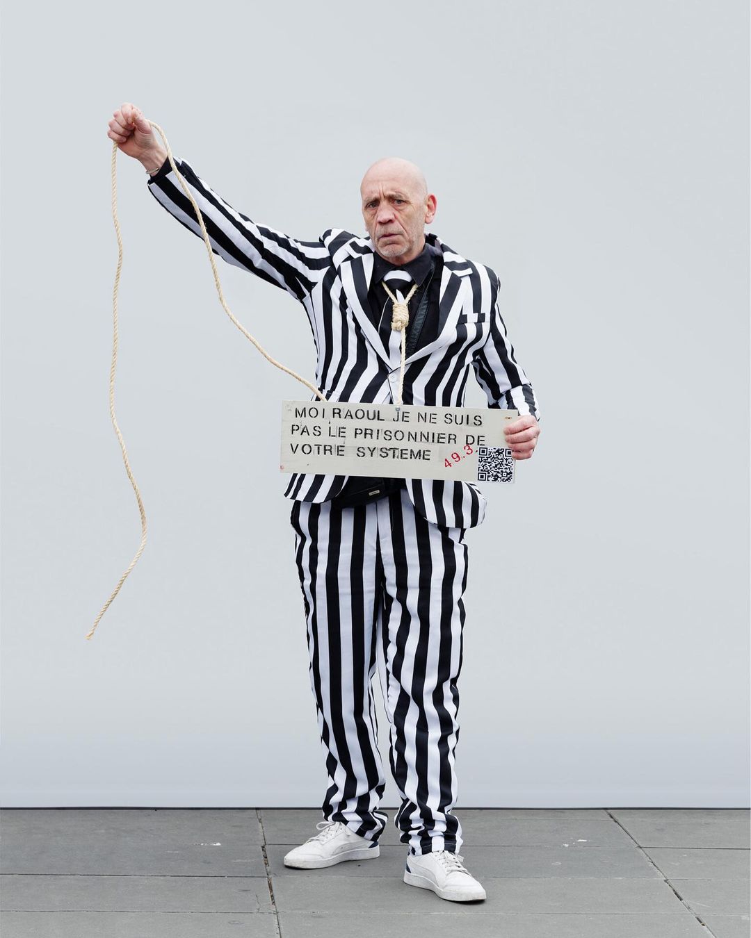 Mature adult in a black-and-white striped suit holds up a sign and rope, standing against a white wall with neutral grey ground. 