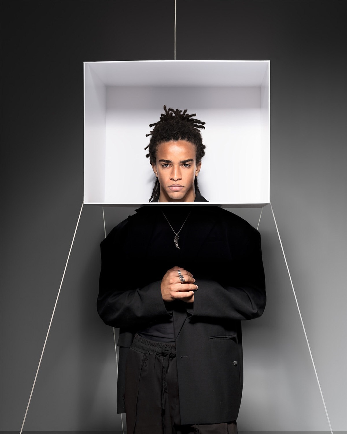 Portrait of an adult male with dreadlocks wearing a black suit. He is positioned behind a white cube suspended in the air.