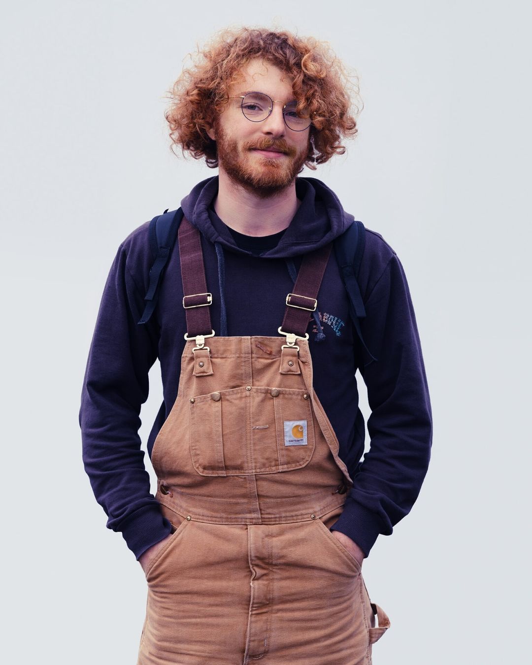 A man with red curly hair, wearing a dark hoodie and tan overalls, stands with hands in pockets against a plain background.