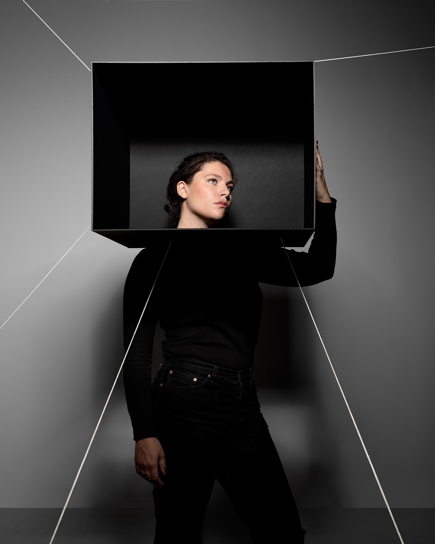 Young woman in black clothing poses inside a black box against a gray background with geometric white lines accentuating the composition.