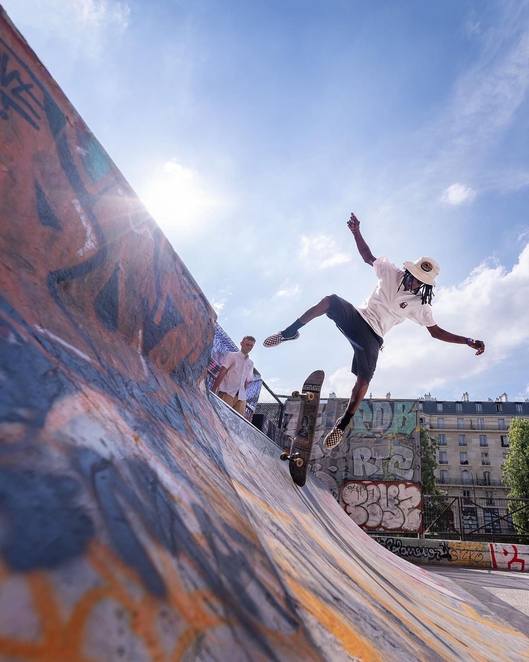 A skateboarder wearing a hat and a white t-shirt is balancing on the skateboard in a skatepark ramp on a sunny day. The buildings are visible in the background.