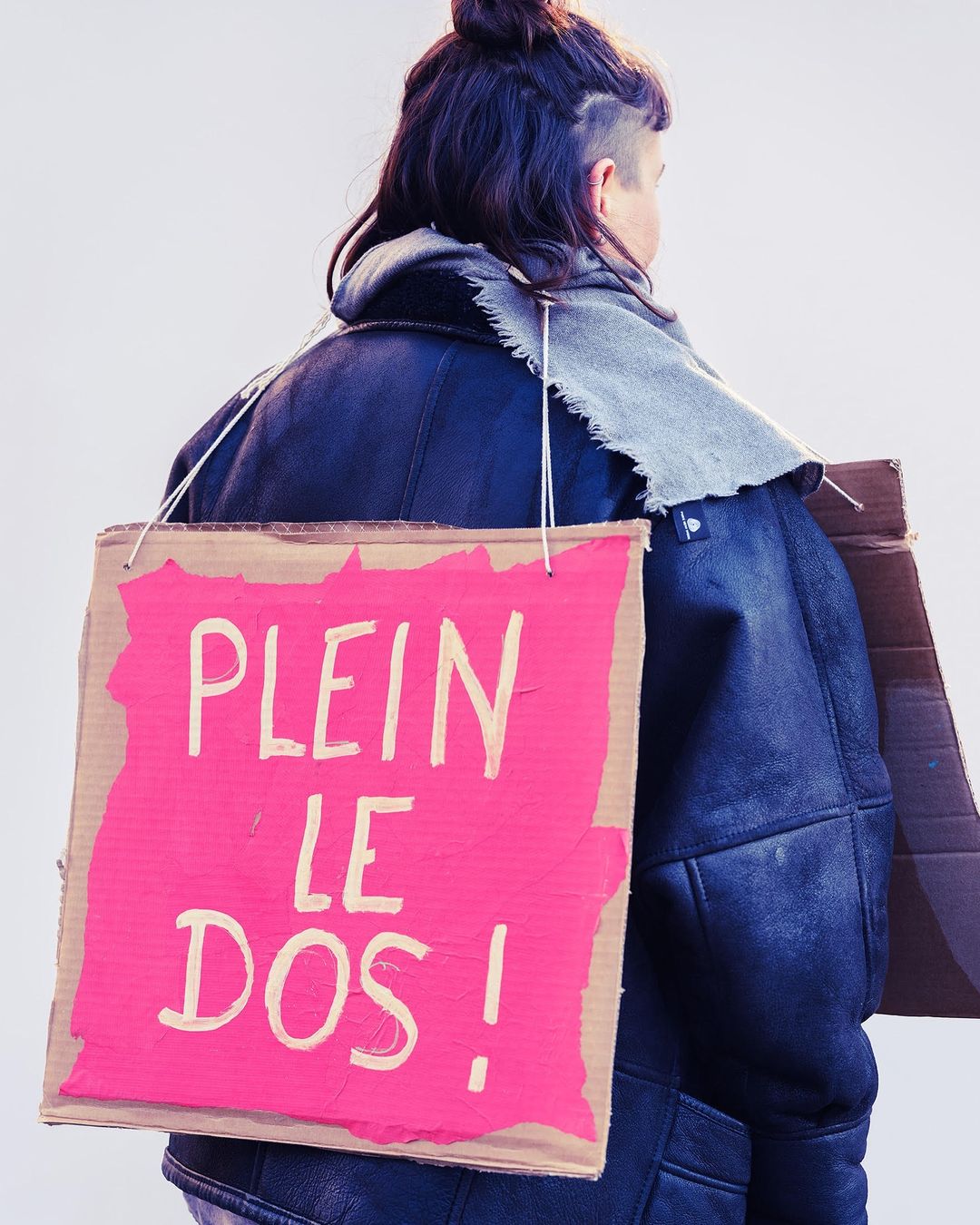 Person from the back wearing a gray scarf and dark blue coat holding a pink sign that says 'PLEIN LE DOS!' hanging on the back.