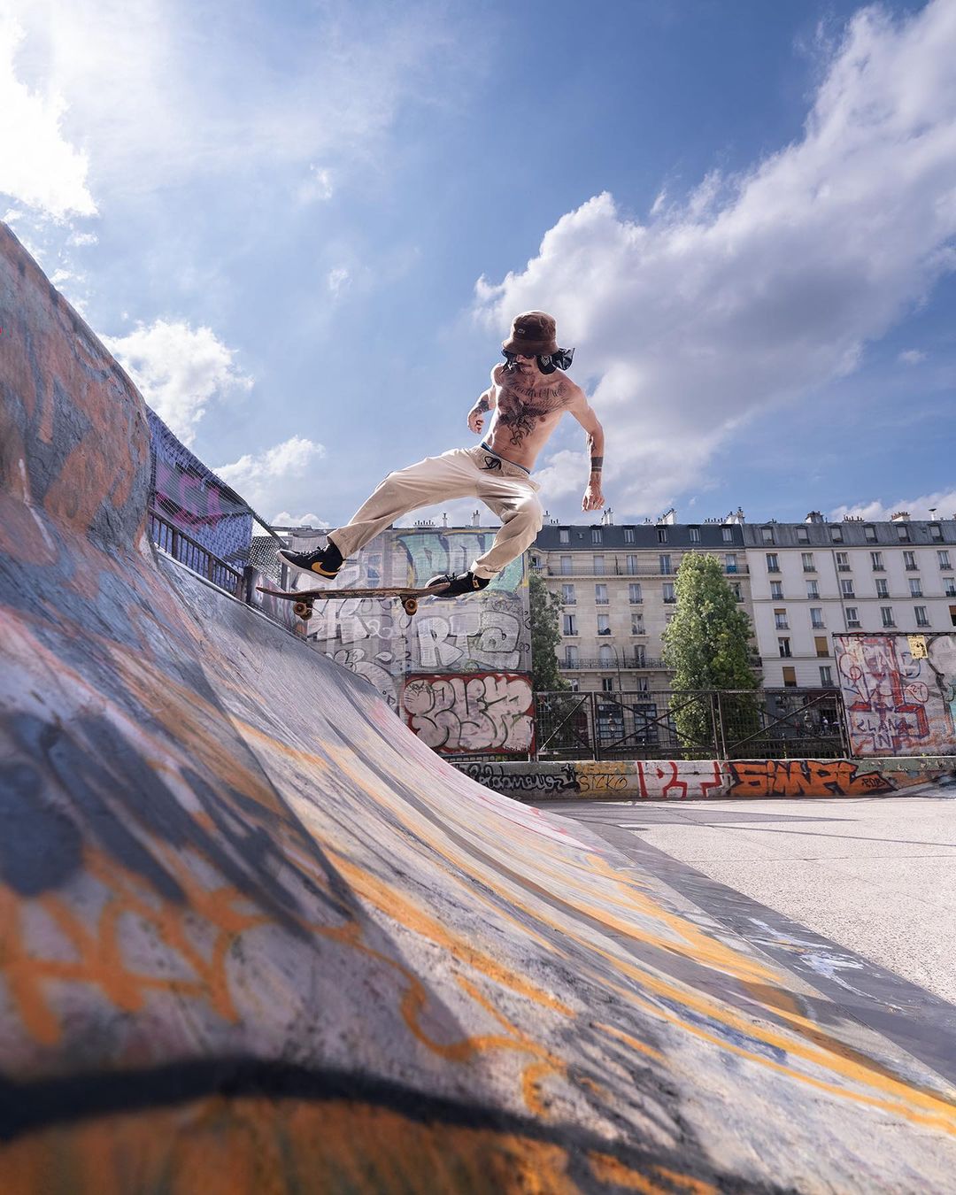 A skateboarder performs a trick on a ramp at an outdoor skatepark under a blue sky with clouds on a sunny day.