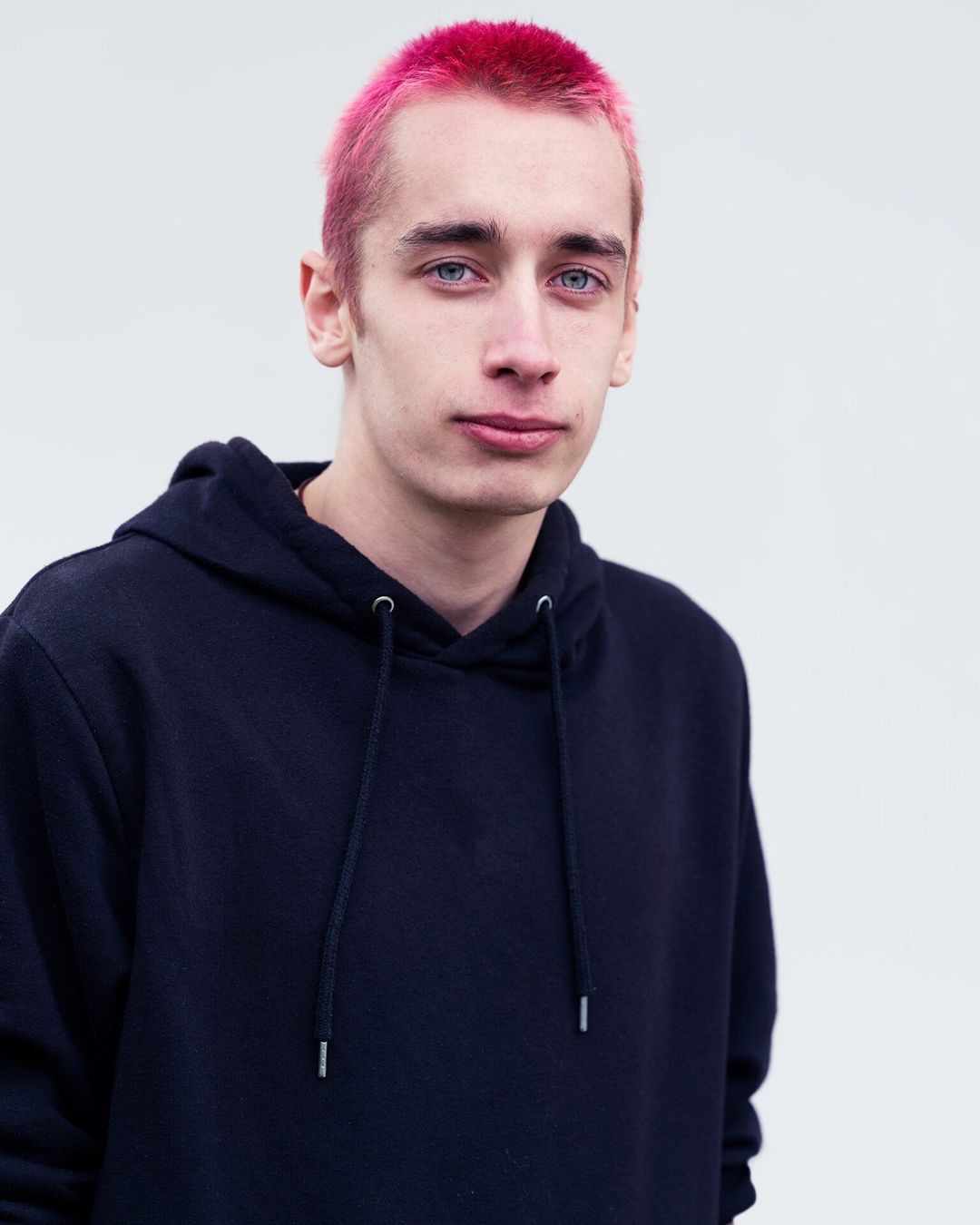 Close-up portrait of a young adult male with dyed pink hair and wearing a black hoodie, set against a plain white background.
