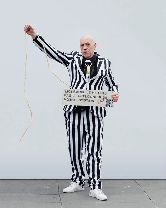 Man wearing black and white striped suit holding a sign with French text and a rope against a plain wall