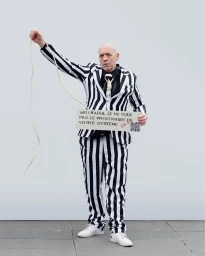 Man wearing black and white striped suit holding a sign with French text and a rope against a plain wall
