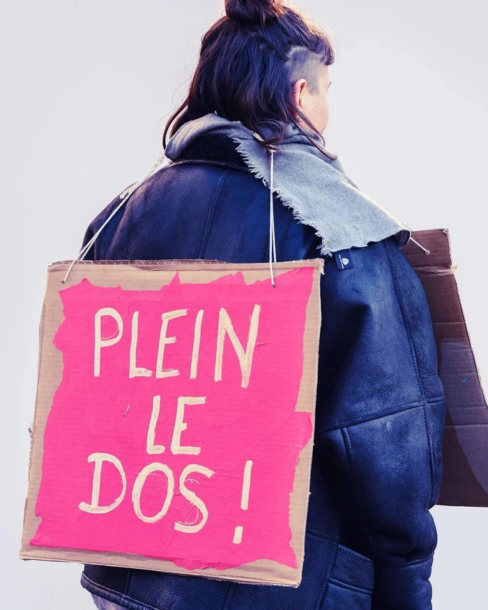 Person seen from behind wearing a dark leather jacket and carrying a cardboard sign on their back with pink painted text reading PLEIN LE DOS!