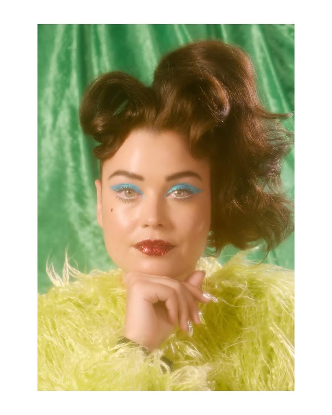 Portrait of a woman with brown hair and bold blue eyeshadow, set against a textured green background, wearing a fuzzy yellow boa.